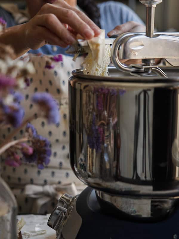 A person grates vegan butter into a stand mixer. They wear a dotted apron, and dried flowers are visible in the foreground.